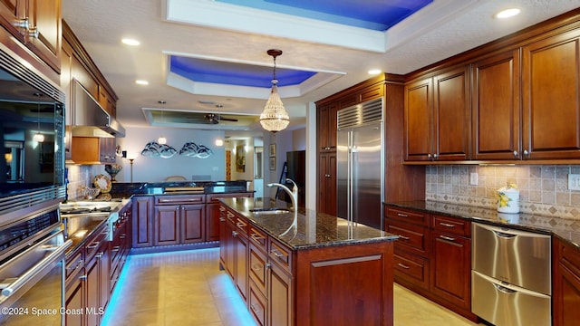 kitchen with backsplash, dark stone countertops, an island with sink, appliances with stainless steel finishes, and a tray ceiling