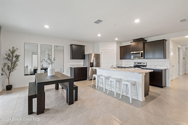 kitchen featuring a kitchen island with sink, dark brown cabinets, light tile patterned floors, and stainless steel appliances