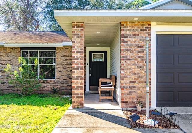 view of exterior entry with a garage, roof with shingles, and brick siding