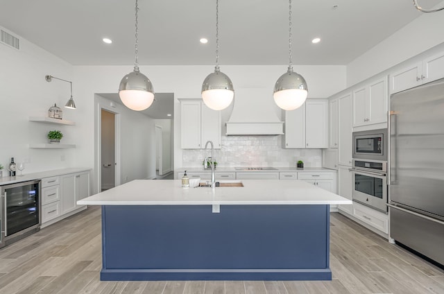 kitchen with beverage cooler, a kitchen island with sink, pendant lighting, built in appliances, and white cabinetry