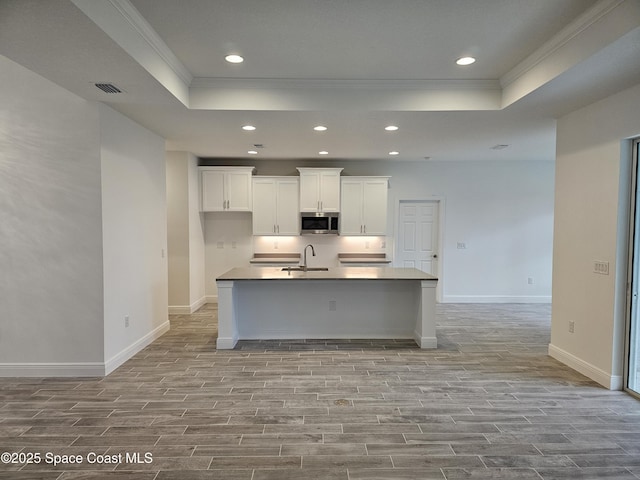 kitchen with a kitchen island with sink, a tray ceiling, ornamental molding, sink, and white cabinets