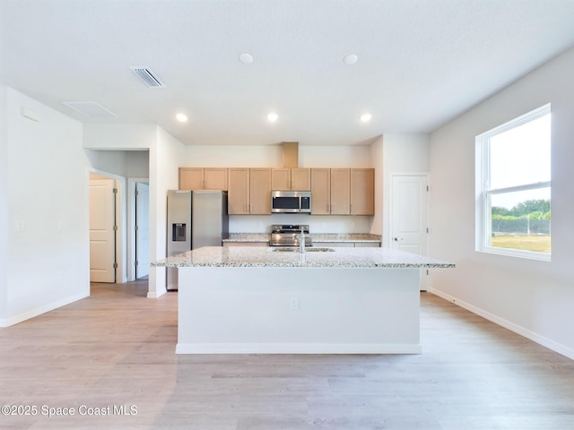 kitchen with light brown cabinets, a center island with sink, light hardwood / wood-style flooring, light stone counters, and stainless steel appliances