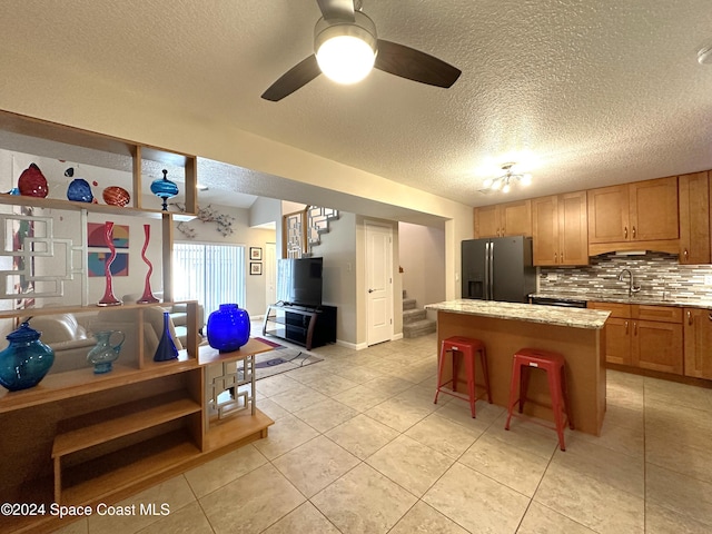 kitchen with refrigerator with ice dispenser, a kitchen island, backsplash, ceiling fan, and a breakfast bar area