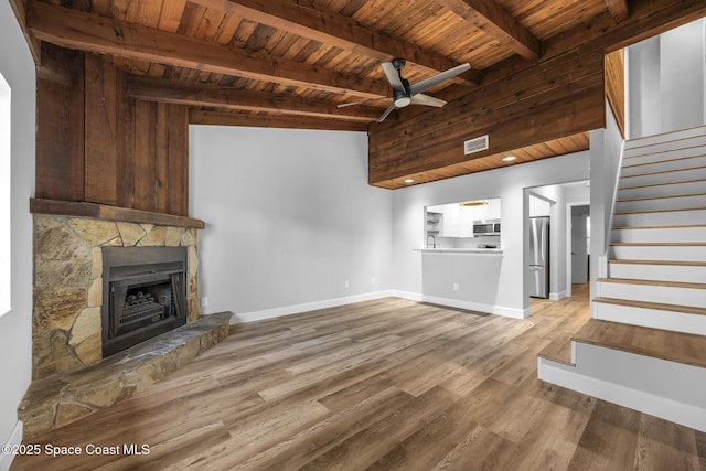 unfurnished living room featuring beam ceiling, ceiling fan, wooden ceiling, a stone fireplace, and light hardwood / wood-style floors