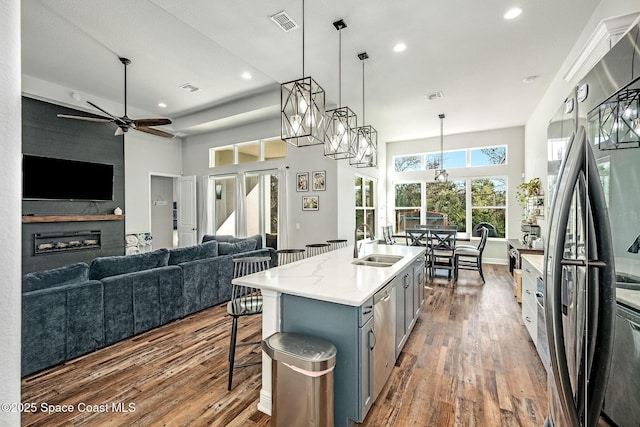 kitchen with ceiling fan, sink, a center island with sink, stainless steel refrigerator, and hanging light fixtures