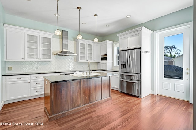 kitchen featuring stainless steel fridge, an island with sink, pendant lighting, wall chimney exhaust hood, and white cabinets