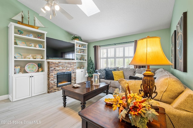 living room featuring ceiling fan, a stone fireplace, light hardwood / wood-style floors, a textured ceiling, and vaulted ceiling with skylight