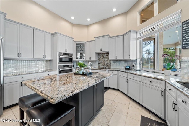 kitchen with stainless steel double oven, a kitchen island with sink, a high ceiling, a kitchen breakfast bar, and backsplash