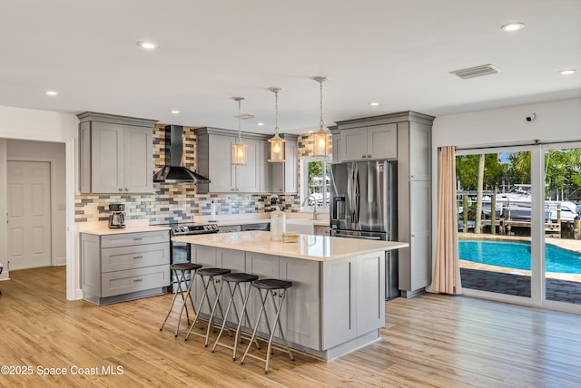 kitchen featuring wall chimney range hood, pendant lighting, a breakfast bar area, appliances with stainless steel finishes, and light wood-type flooring