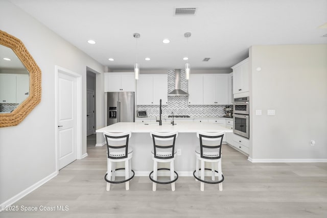 kitchen with pendant lighting, a kitchen island with sink, wall chimney range hood, white cabinetry, and stainless steel appliances