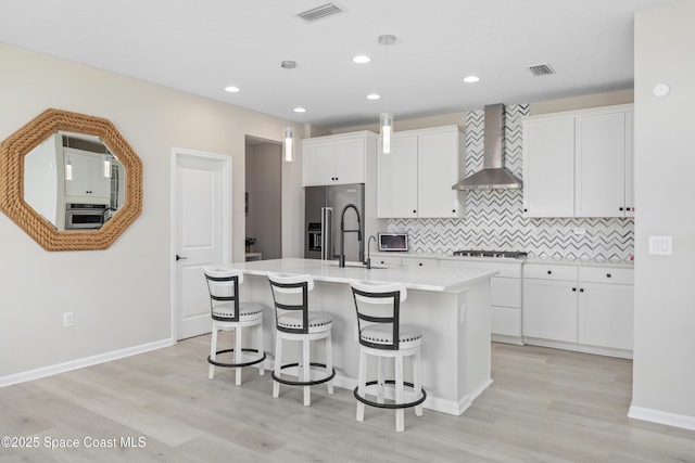 kitchen with pendant lighting, a center island with sink, wall chimney range hood, appliances with stainless steel finishes, and white cabinetry