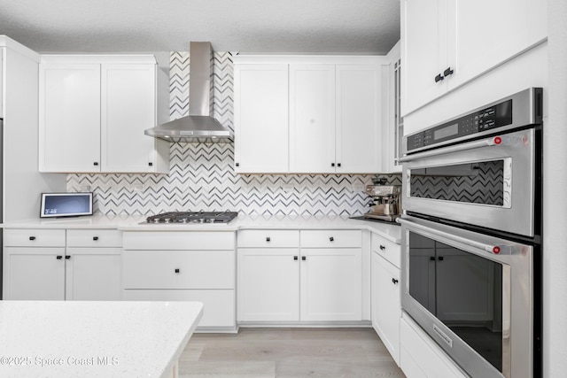 kitchen featuring stainless steel appliances, wall chimney range hood, a textured ceiling, decorative backsplash, and white cabinets