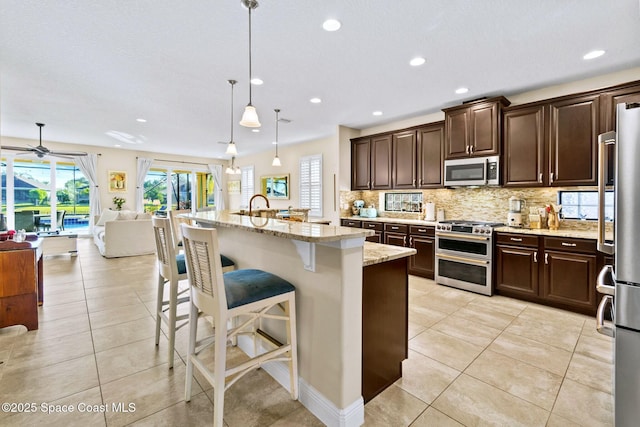 kitchen with light stone countertops, appliances with stainless steel finishes, a kitchen breakfast bar, a kitchen island with sink, and hanging light fixtures