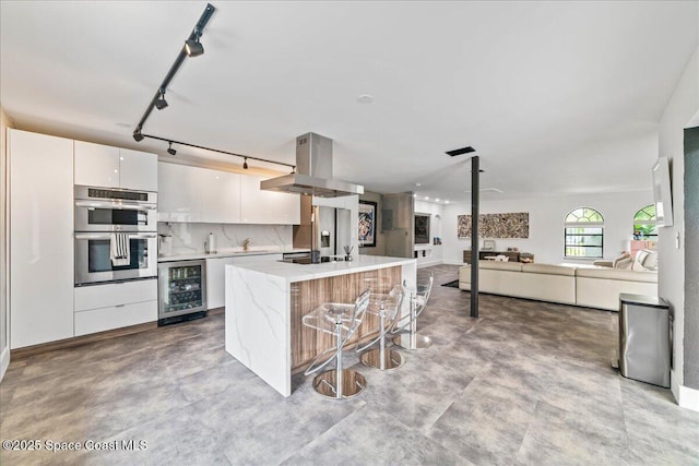 kitchen featuring stainless steel appliances, island range hood, a kitchen island with sink, beverage cooler, and white cabinetry