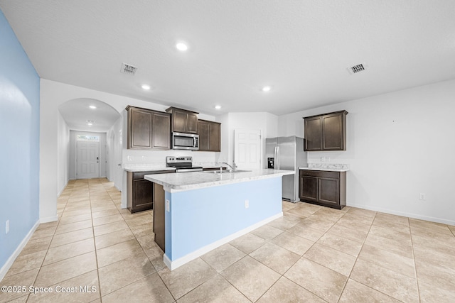 kitchen with dark brown cabinets, a textured ceiling, stainless steel appliances, light tile patterned floors, and an island with sink