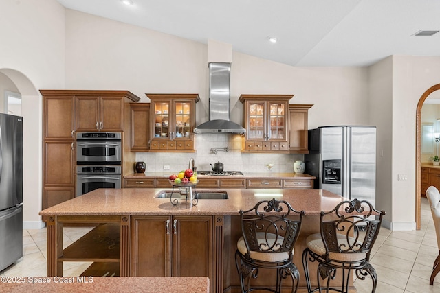 kitchen featuring light tile patterned flooring, appliances with stainless steel finishes, an island with sink, decorative backsplash, and wall chimney exhaust hood