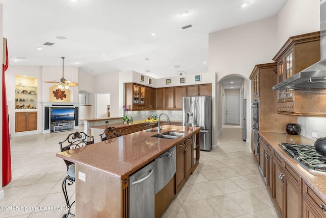 kitchen with a kitchen bar, sink, appliances with stainless steel finishes, a kitchen island with sink, and wall chimney range hood