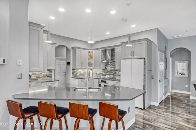 kitchen featuring hanging light fixtures, wall chimney range hood, white refrigerator, kitchen peninsula, and a breakfast bar area