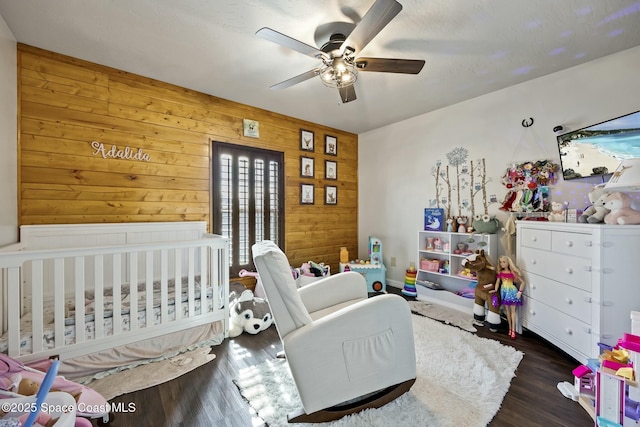 bedroom featuring ceiling fan, wood walls, dark hardwood / wood-style flooring, and a nursery area