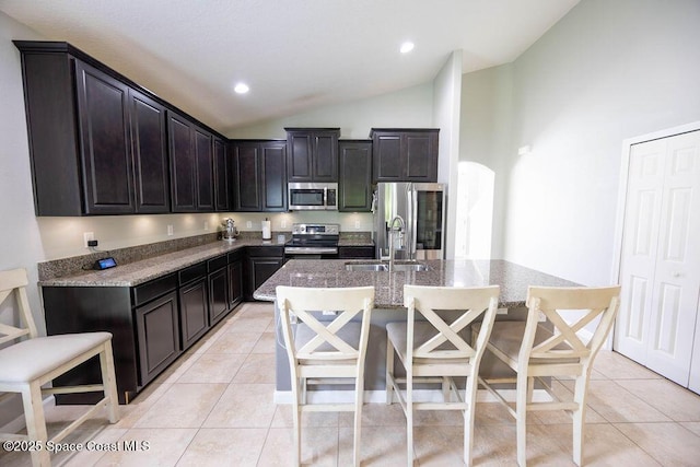 kitchen with vaulted ceiling, light tile patterned floors, an island with sink, light stone countertops, and appliances with stainless steel finishes