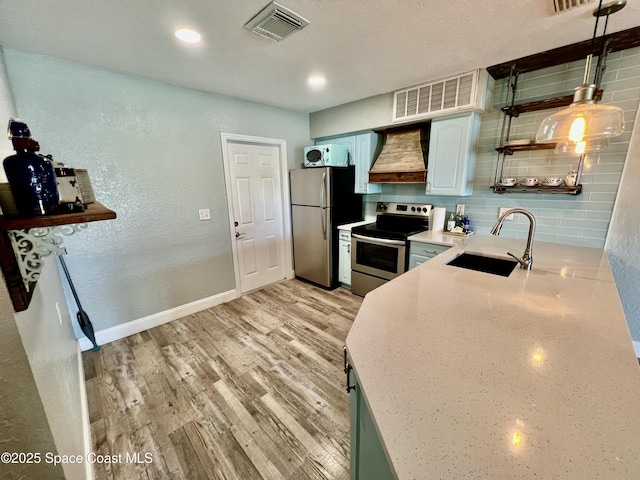 kitchen featuring sink, custom exhaust hood, hanging light fixtures, light wood-type flooring, and appliances with stainless steel finishes