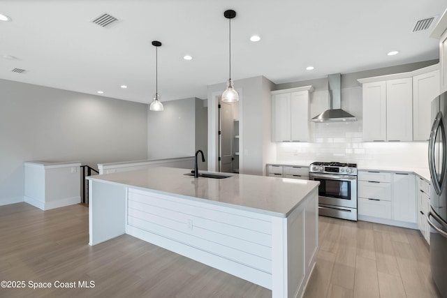 kitchen with white cabinetry, gas range, wall chimney range hood, sink, and an island with sink