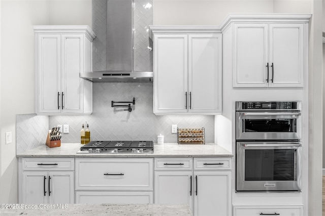 kitchen featuring backsplash, white cabinetry, light stone counters, wall chimney range hood, and stainless steel appliances