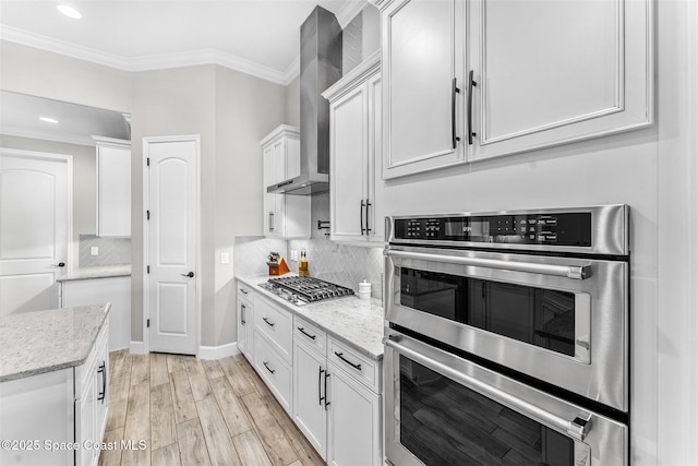 kitchen with light hardwood / wood-style flooring, appliances with stainless steel finishes, white cabinets, light stone counters, and wall chimney range hood