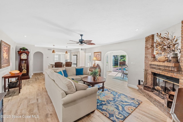 living room featuring arched walkways, a fireplace, a ceiling fan, baseboards, and light wood-style floors