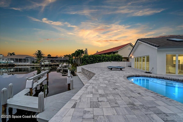 pool at dusk with a dock, a water view, and a patio