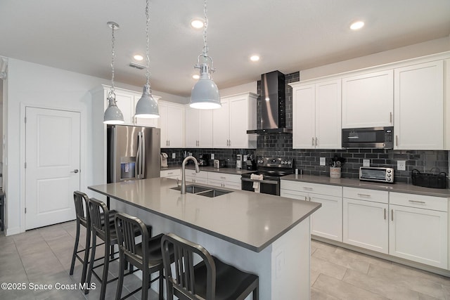 kitchen featuring an island with sink, appliances with stainless steel finishes, hanging light fixtures, wall chimney range hood, and sink