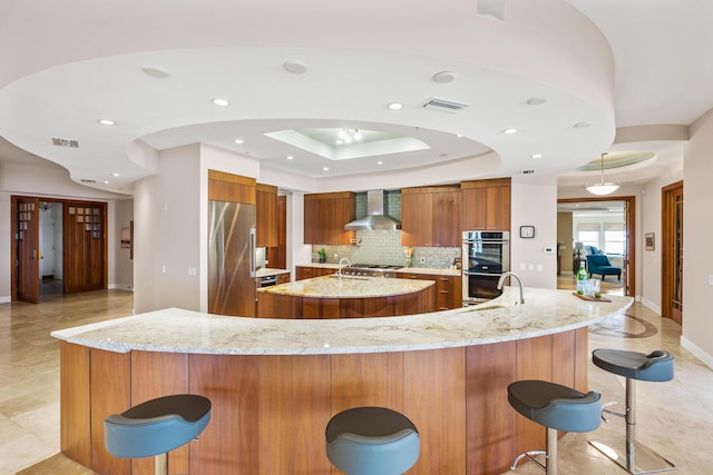 kitchen featuring kitchen peninsula, a tray ceiling, appliances with stainless steel finishes, a breakfast bar area, and wall chimney exhaust hood