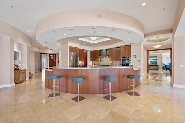 kitchen with wall chimney range hood, stainless steel appliances, tasteful backsplash, a raised ceiling, and a breakfast bar