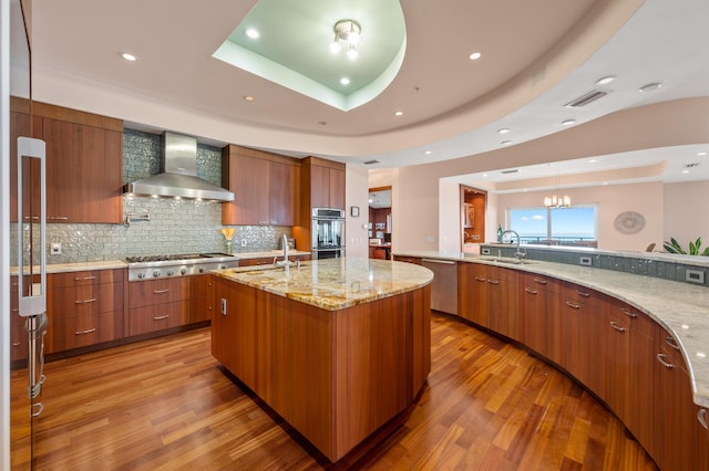 kitchen with appliances with stainless steel finishes, wall chimney exhaust hood, sink, a raised ceiling, and a center island with sink