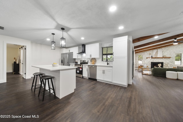 kitchen with pendant lighting, appliances with stainless steel finishes, a stone fireplace, white cabinets, and wall chimney exhaust hood