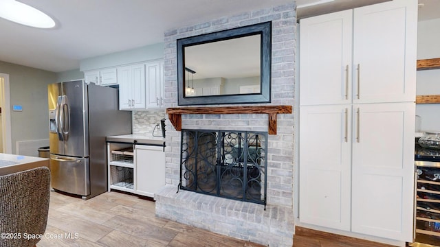 kitchen with stainless steel refrigerator with ice dispenser, white cabinetry, light hardwood / wood-style flooring, a brick fireplace, and backsplash