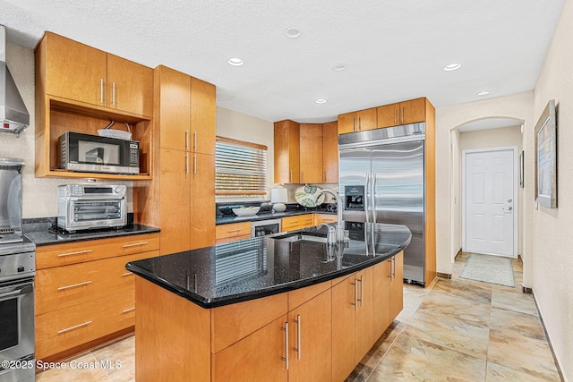 kitchen with sink, dark stone counters, a kitchen island with sink, stainless steel appliances, and wall chimney exhaust hood