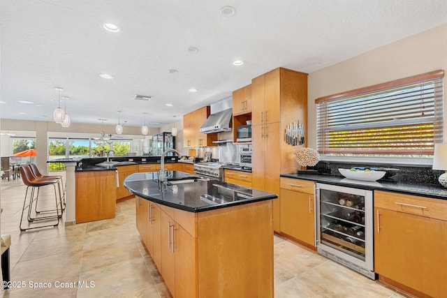 kitchen with wall chimney range hood, sink, appliances with stainless steel finishes, an island with sink, and beverage cooler