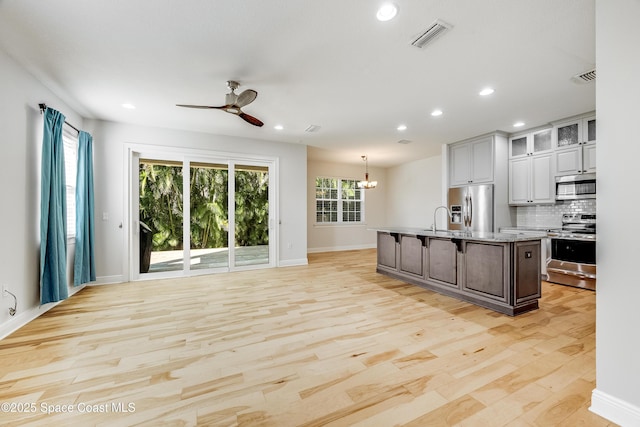 kitchen with stainless steel appliances, tasteful backsplash, a center island with sink, decorative light fixtures, and light wood-type flooring