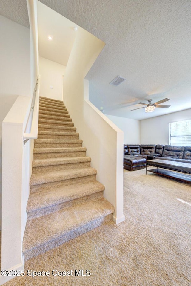 stairs with ceiling fan, a textured ceiling, and carpet