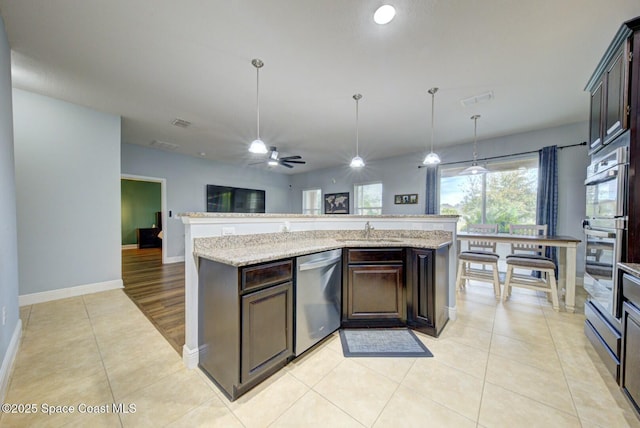 kitchen with pendant lighting, dark brown cabinets, stainless steel appliances, light stone countertops, and a center island with sink