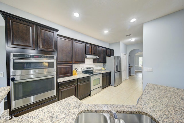 kitchen featuring appliances with stainless steel finishes, backsplash, light tile patterned floors, light stone countertops, and dark brown cabinets