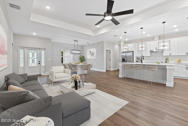 living room with light wood-type flooring, ceiling fan with notable chandelier, and a tray ceiling