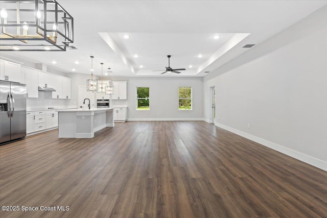 unfurnished living room with sink, ceiling fan with notable chandelier, a tray ceiling, and dark hardwood / wood-style flooring