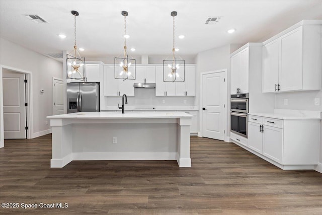 kitchen featuring appliances with stainless steel finishes, decorative light fixtures, dark wood-type flooring, white cabinetry, and an island with sink