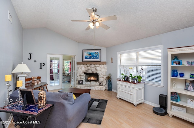 living room featuring vaulted ceiling, a stone fireplace, light wood-type flooring, ceiling fan, and a textured ceiling