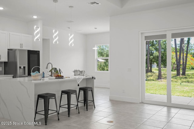 kitchen featuring stainless steel refrigerator with ice dispenser, hanging light fixtures, white cabinetry, light stone countertops, and a breakfast bar