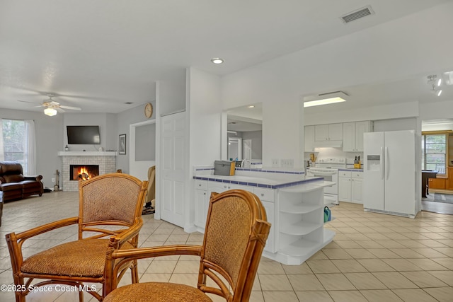 kitchen with white appliances, a brick fireplace, light tile patterned floors, white cabinets, and plenty of natural light