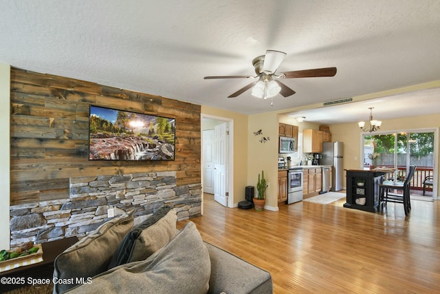 living room with ceiling fan with notable chandelier, a textured ceiling, light wood-type flooring, and wood walls