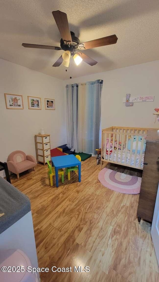 bedroom with ceiling fan, a nursery area, a textured ceiling, and light wood-type flooring